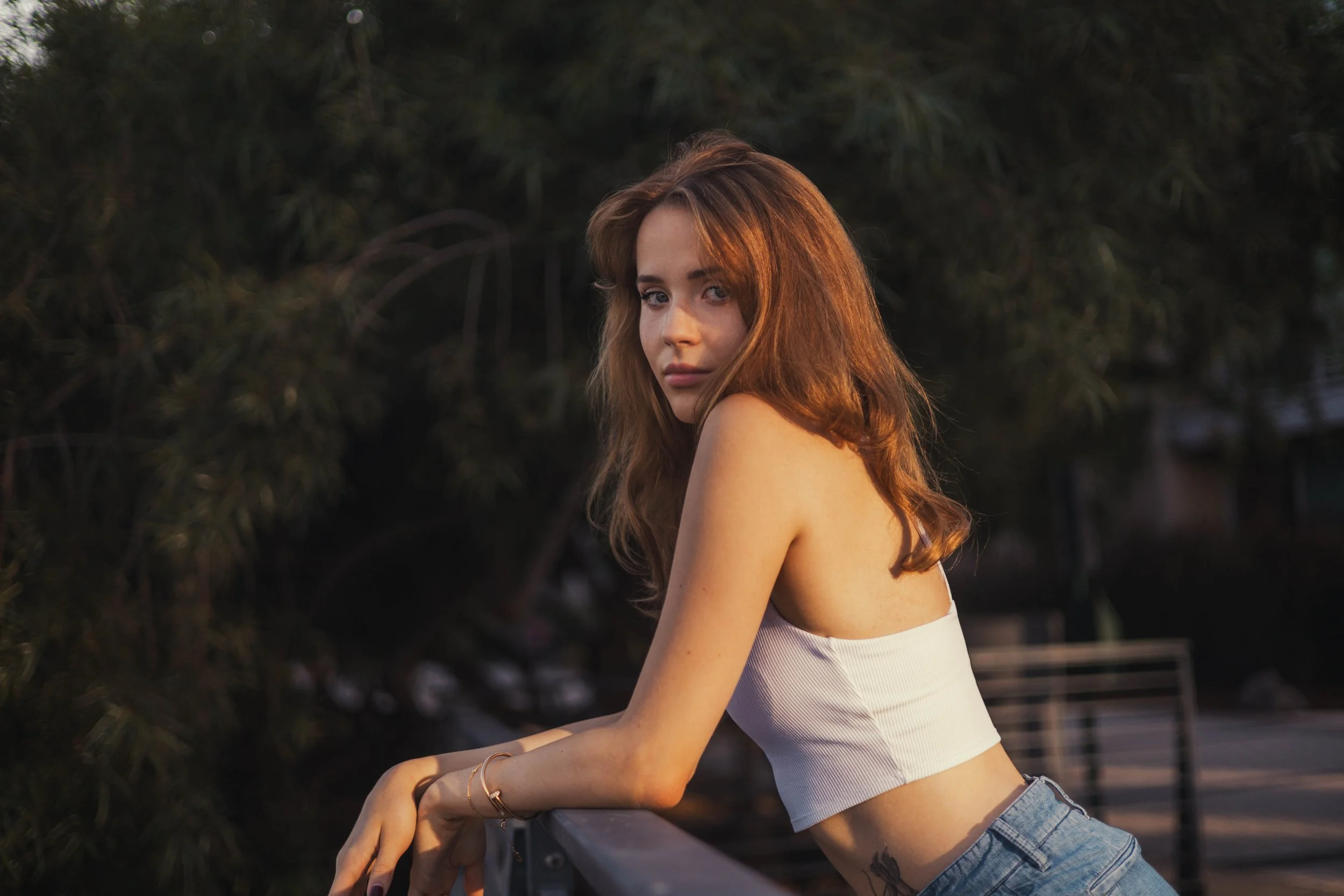 A young woman with long, wavy, reddish-brown hair leaning on a railing outdoors during golden hour, wearing a white crop top and jeans.
