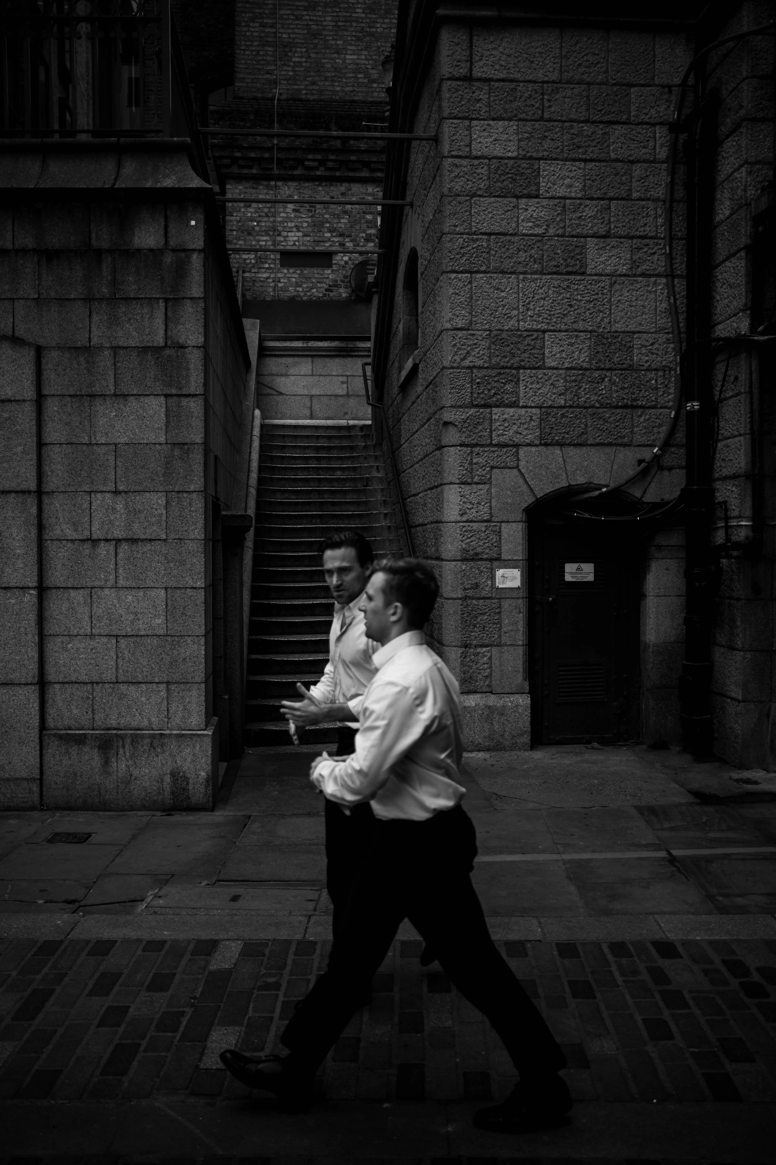 Two men dressed in white shirts and dark pants walking in an alleyway, with stone buildings and a staircase in the background, captured in black and white.