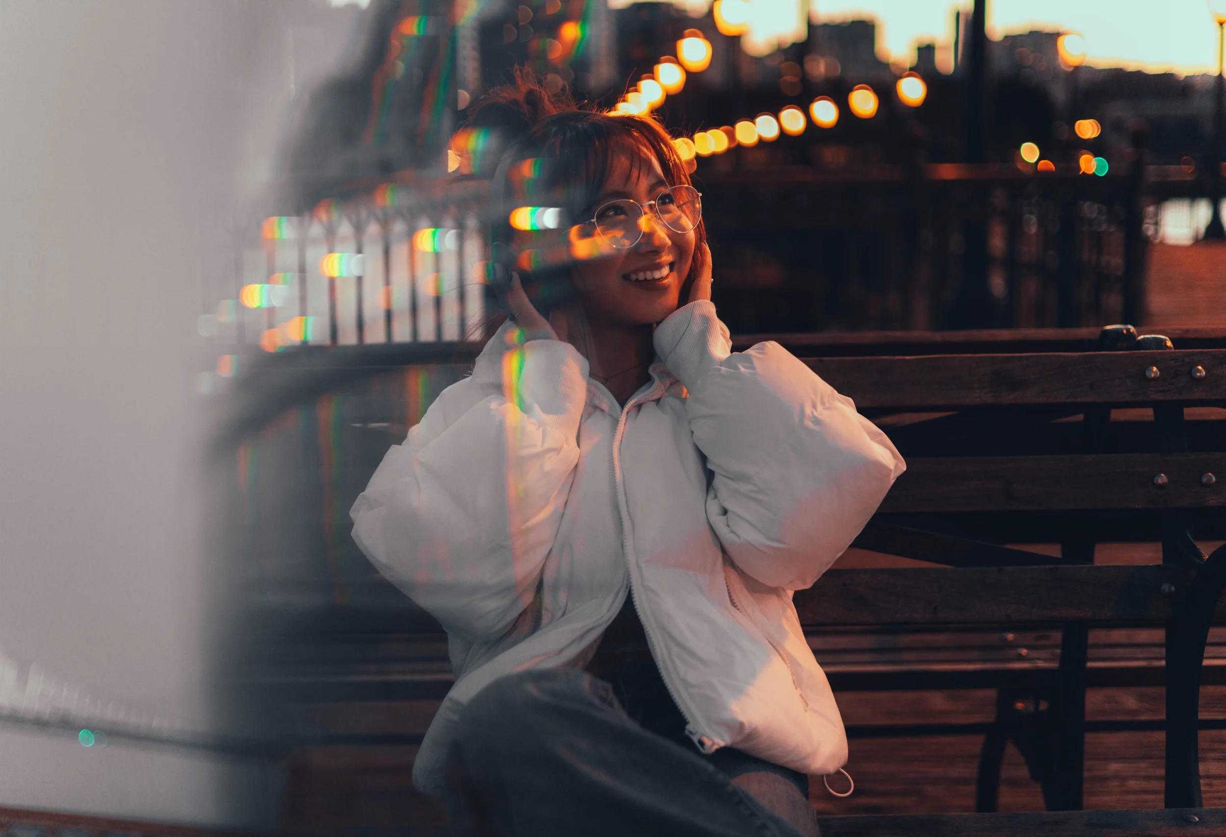 A young woman with glasses smiling and listening to music with headphones on, sitting on a wooden bench outdoors during sunset, with city lights in the background.