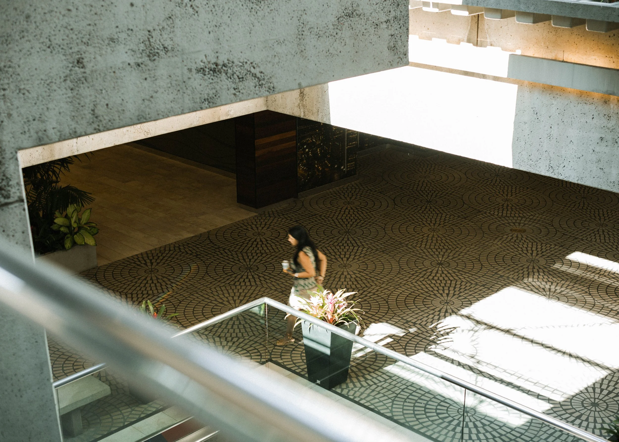 A woman with long dark hair walking through a modern building lobby, holding a drink, with patterned carpet, potted plants, and architectural concrete surfaces.