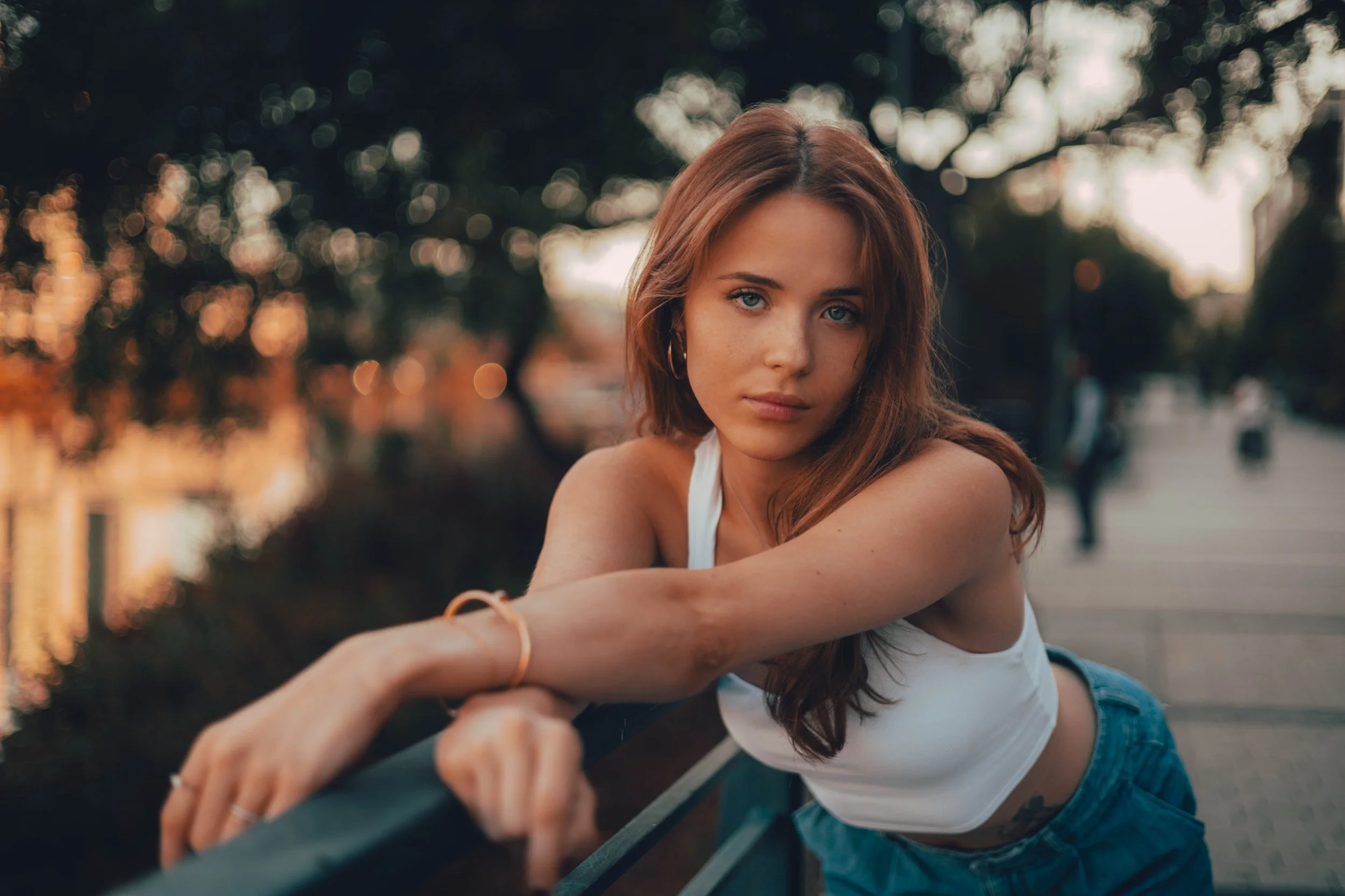 A young woman with red hair and blue eyes leaning on a railing outdoors during sunset or dusk.