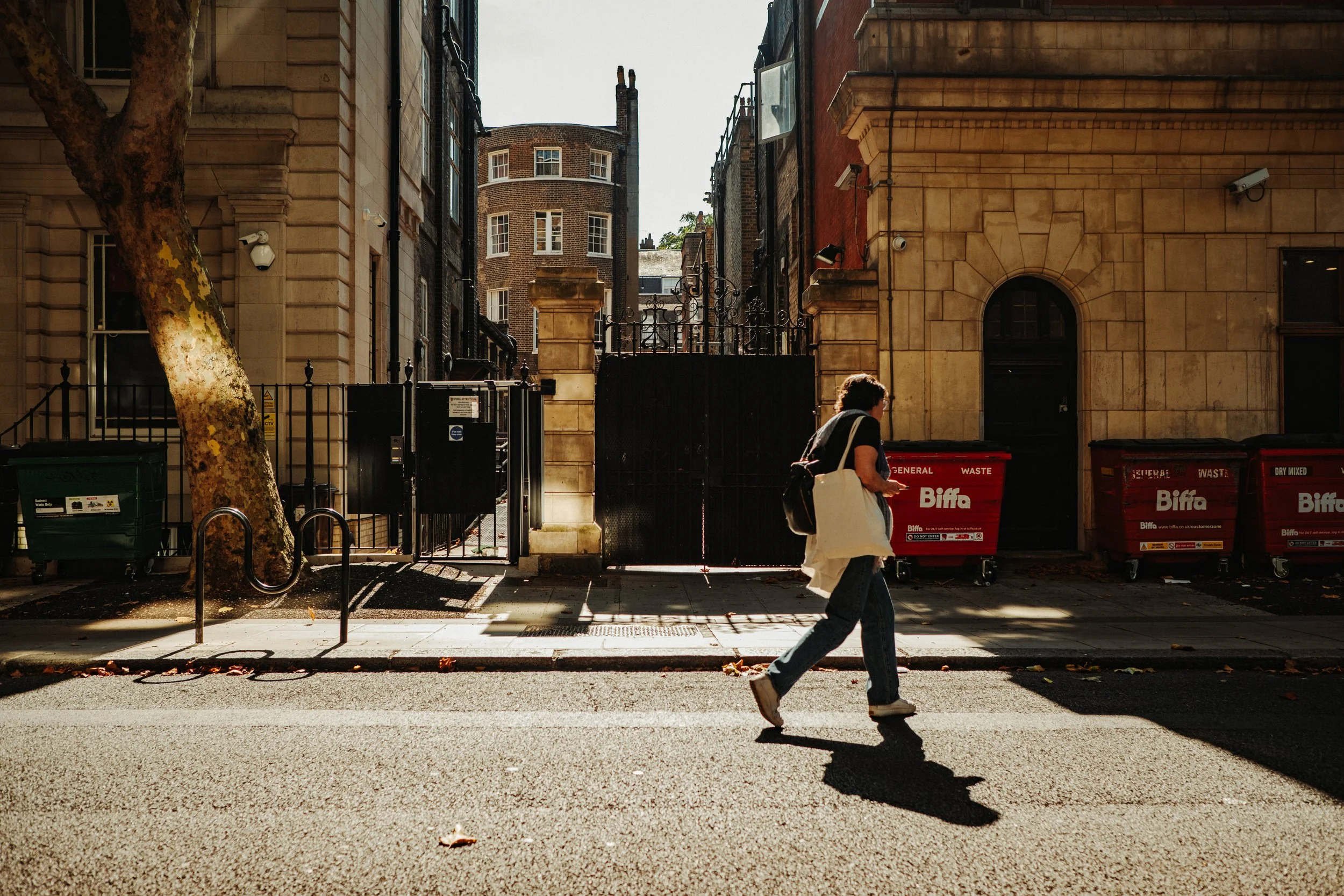 A person walking across a city street, carrying a tote bag and wearing sunglasses, with historic buildings, dumpsters, and a large tree in the background.