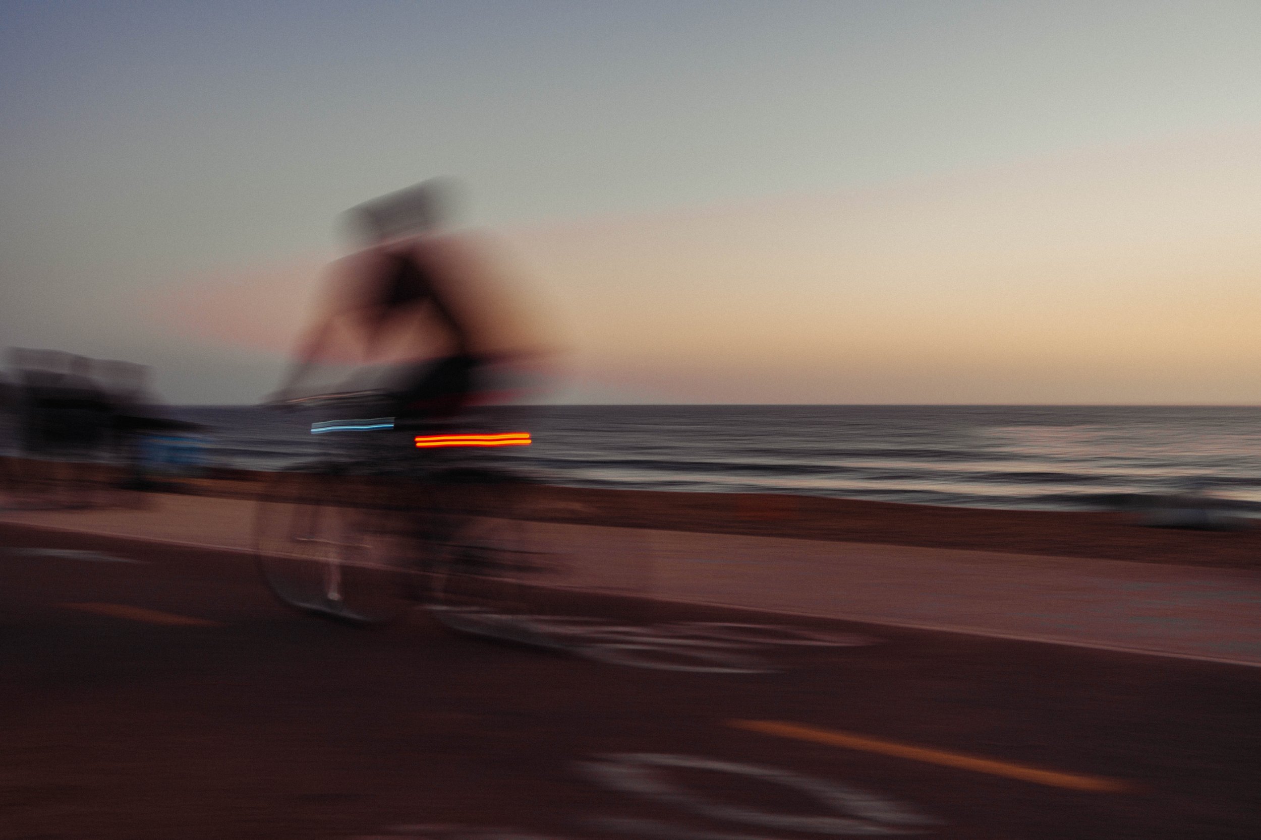 Blurry photo of a cyclist riding on a road by the ocean during sunset with a pastel-colored sky.