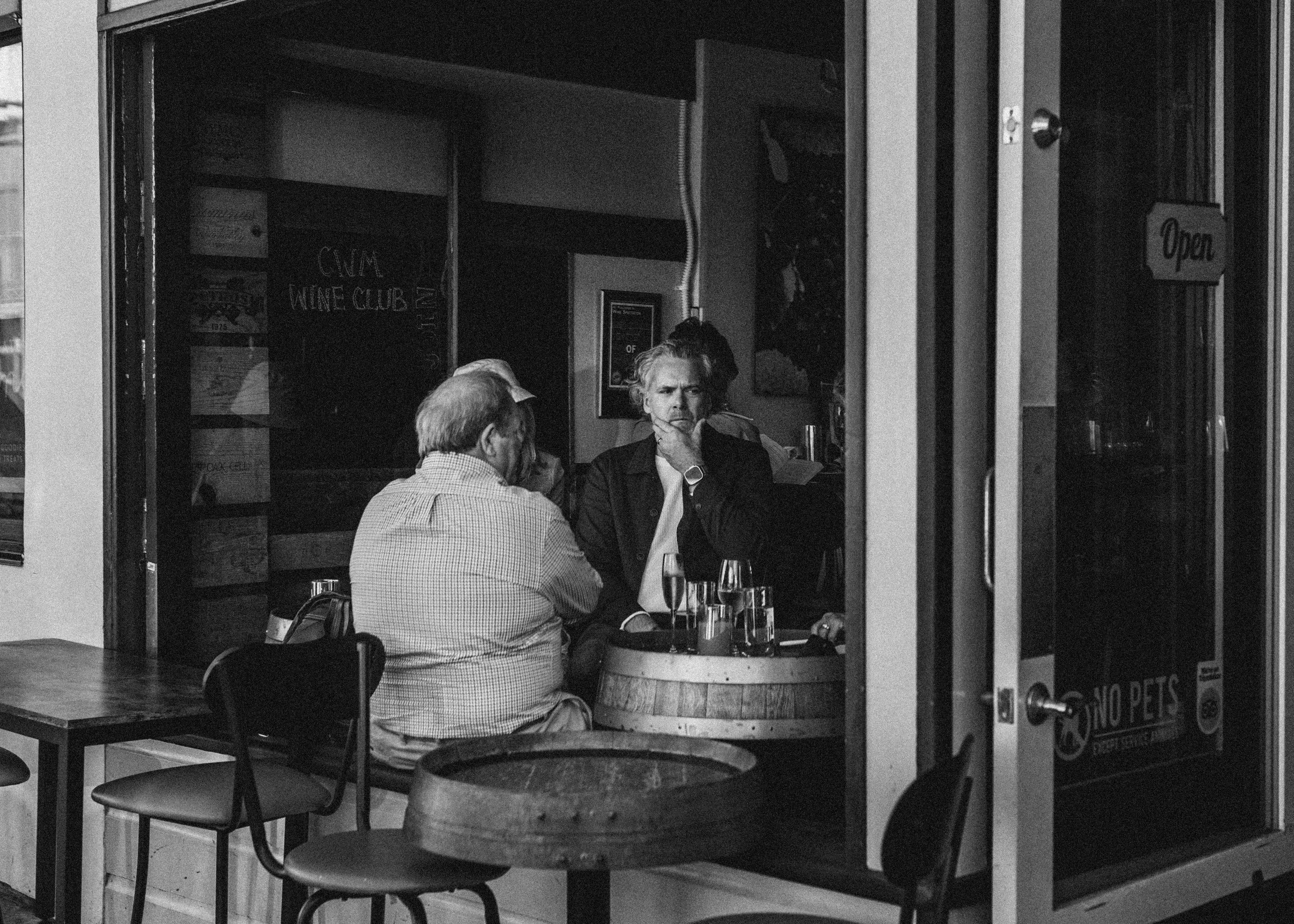 Two men sitting at a table in a cafe, one with a beard and glasses, and the other with longer hair, deep in conversation, with drinks on the table, seen through a large window.