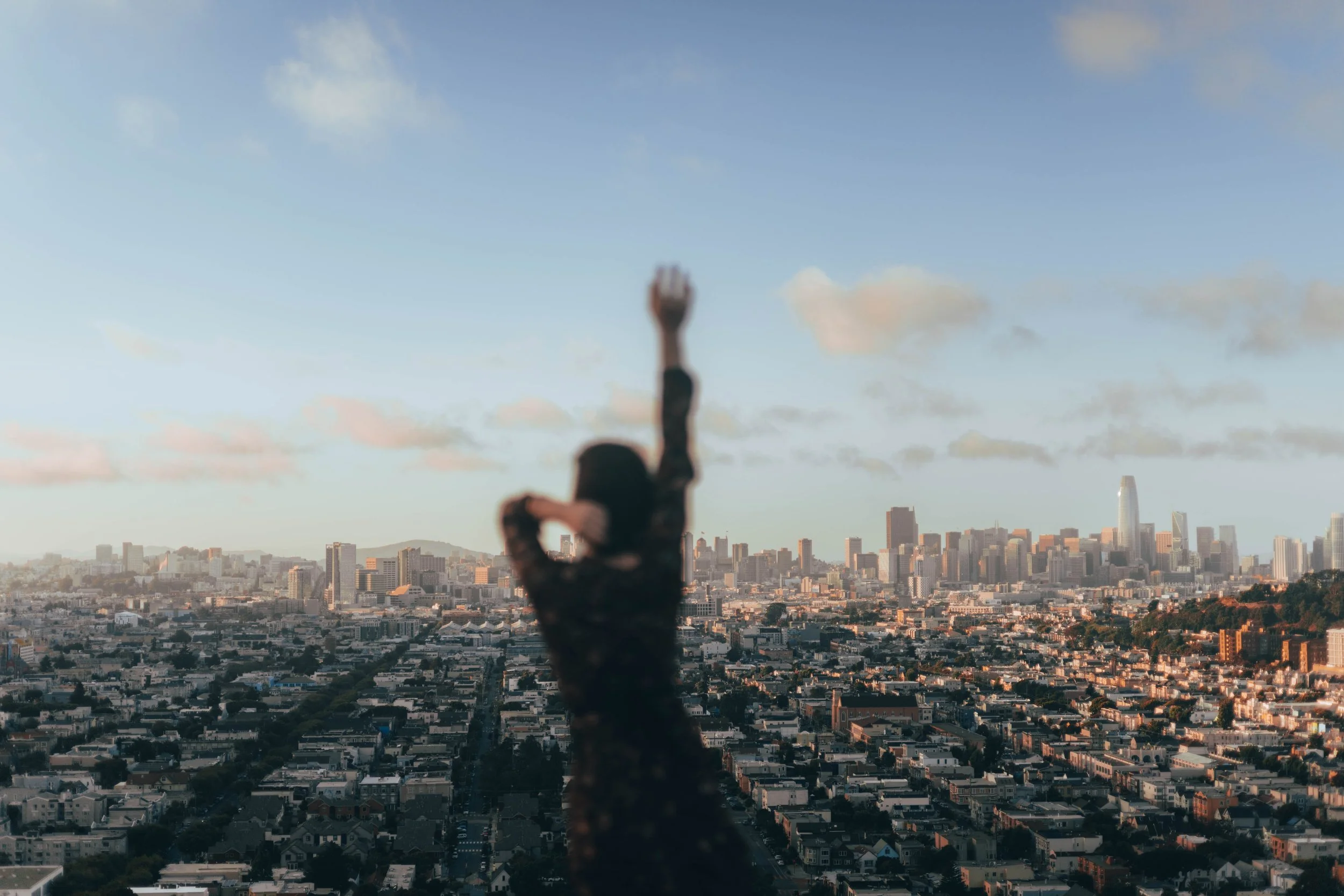 A blurry woman with dark hair standing on a rooftop with San Francisco city skyline in the background, raising one arm and touching her head.
