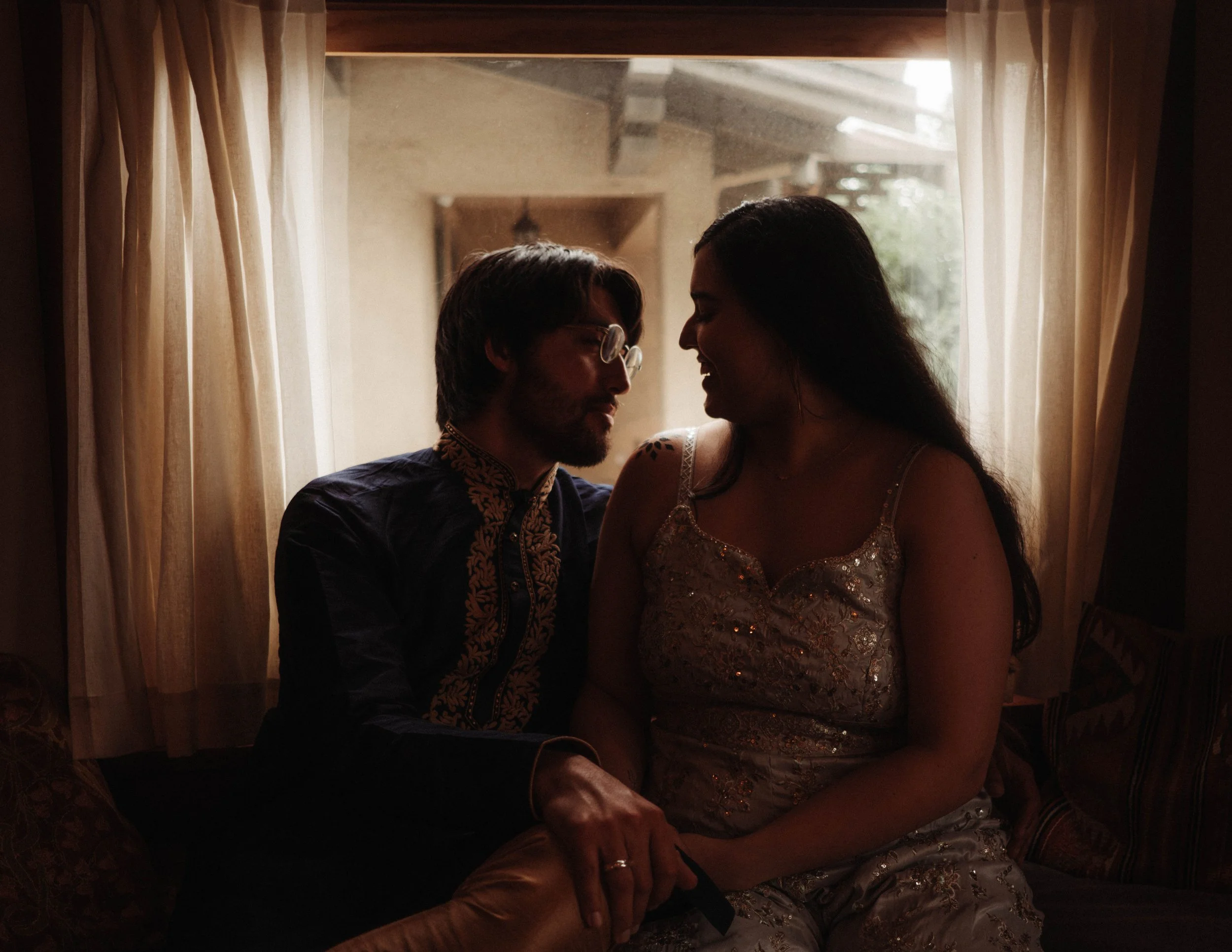 A man and woman sitting close together in a dimly lit room, smiling and leaning towards each other. The background shows a window with curtains and outdoor greenery.