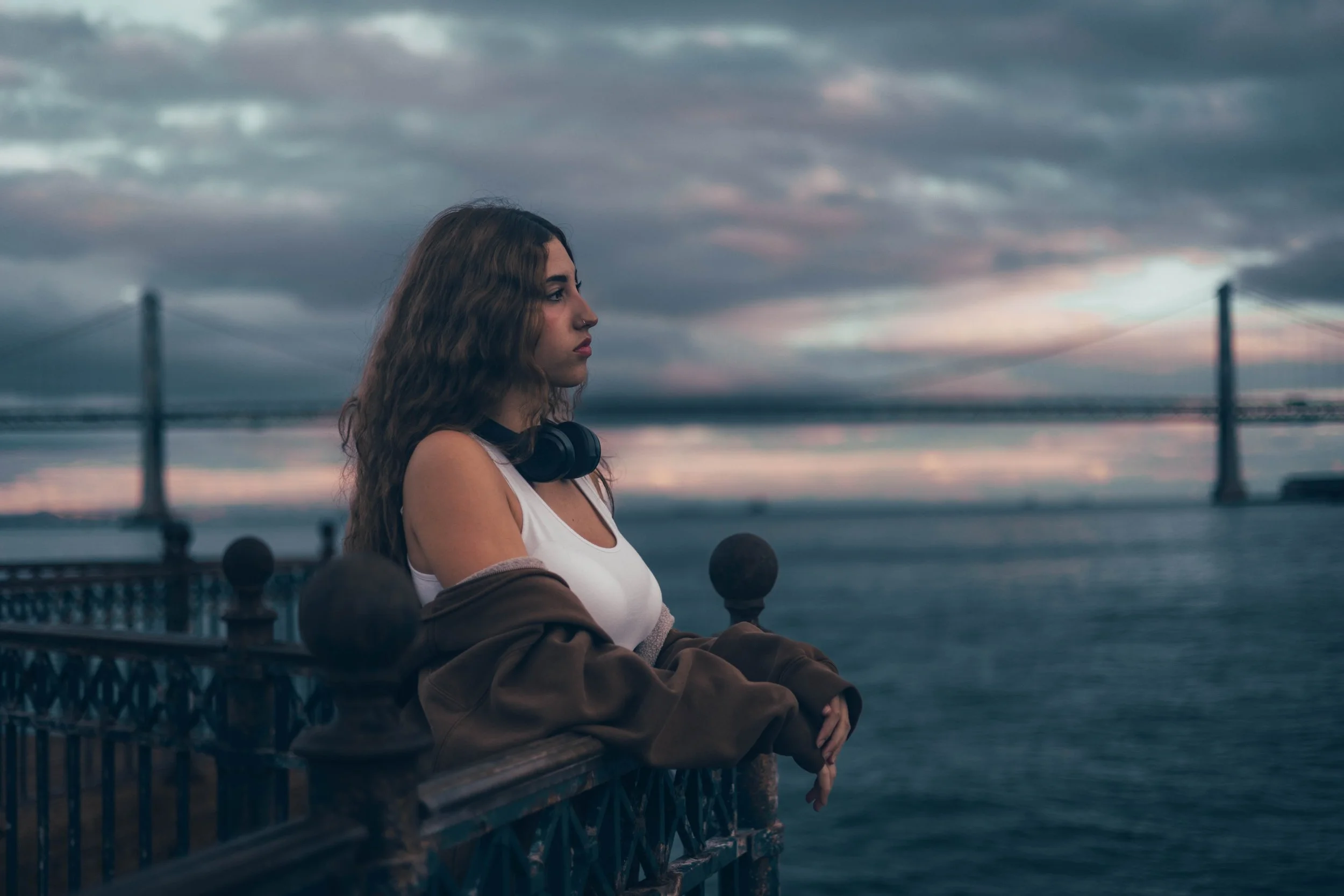 A young woman with long wavy hair stands on a bridge overlooking the water at dusk, wearing a white top, a brown jacket draped over her shoulders, and black headphones around her neck, with a large bridge in the background under a cloudy sky.