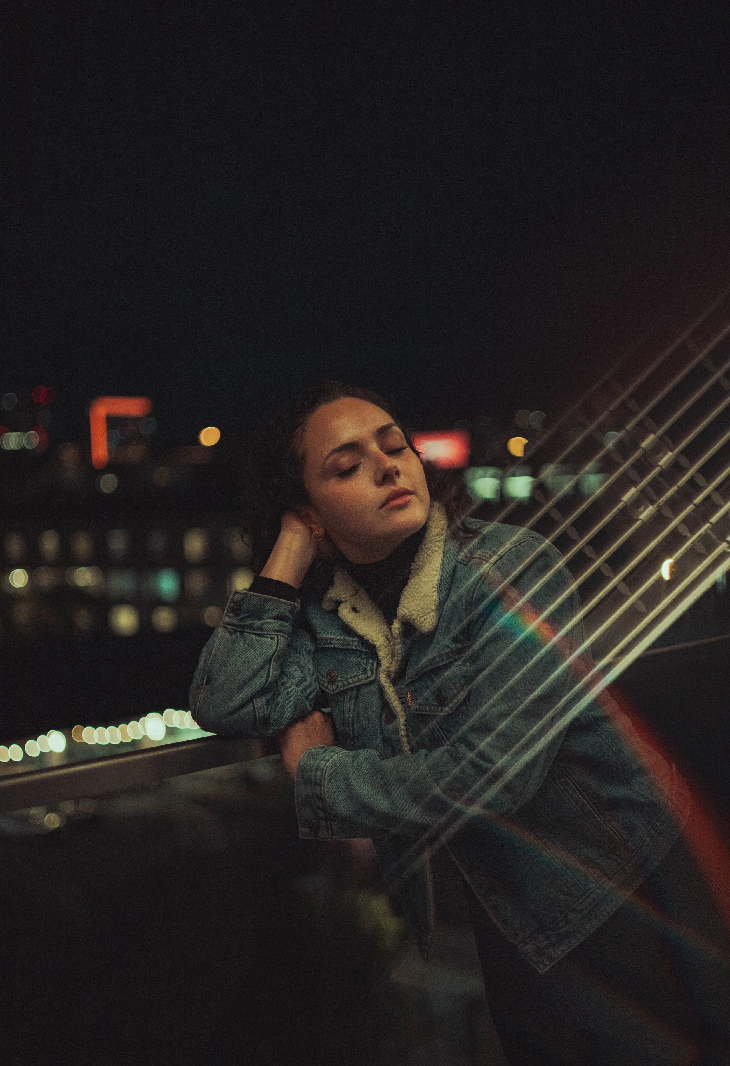 A young woman at night near a railing, resting her head on her hand with her eyes closed, wearing a denim jacket and a black turtleneck, city lights in the background.