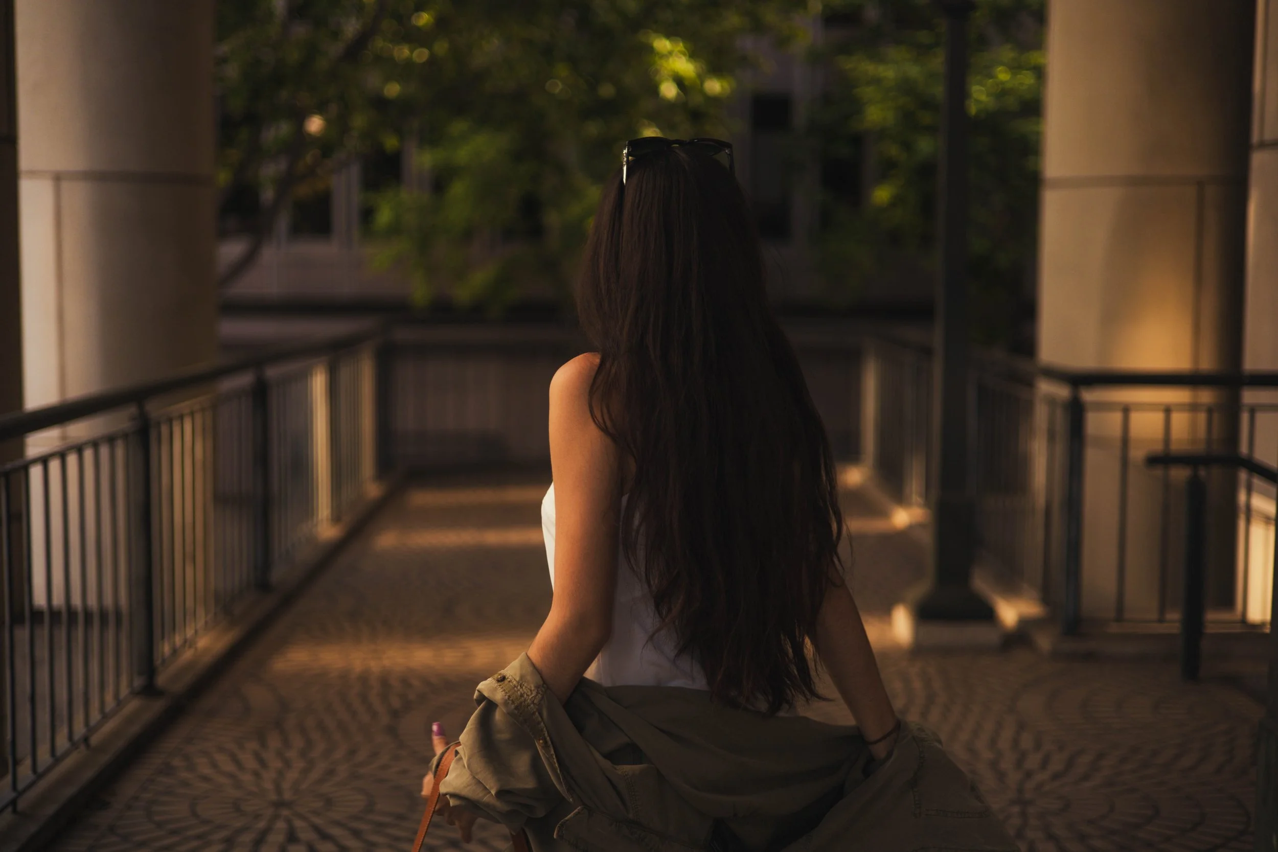 A woman with long dark hair wearing sunglasses and a white top, sitting on her knees on a patterned outdoor pavement at dusk, with greenery and building windows in the background.