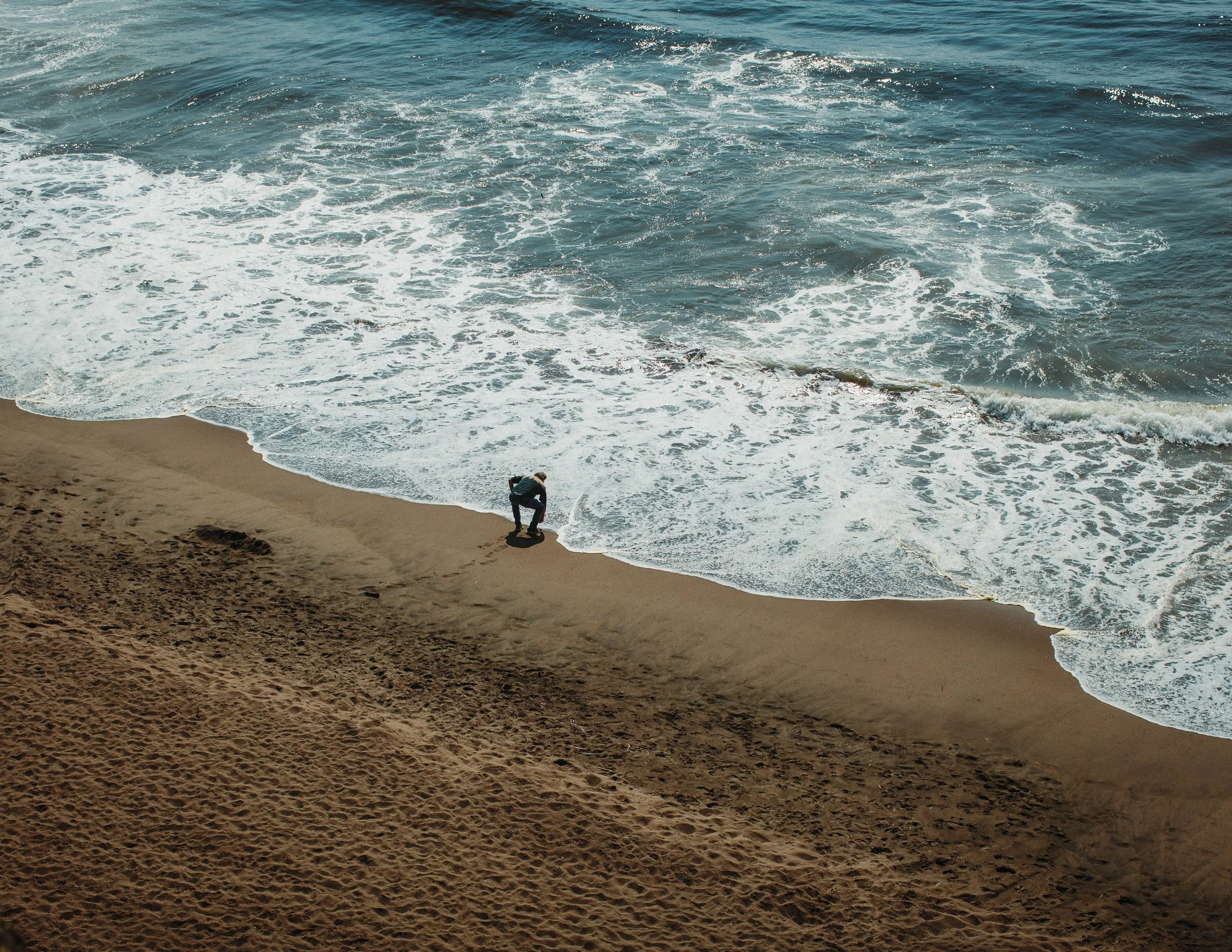 A person on a skateboard near the shoreline of an ocean beach, with waves crashing onto the sand.