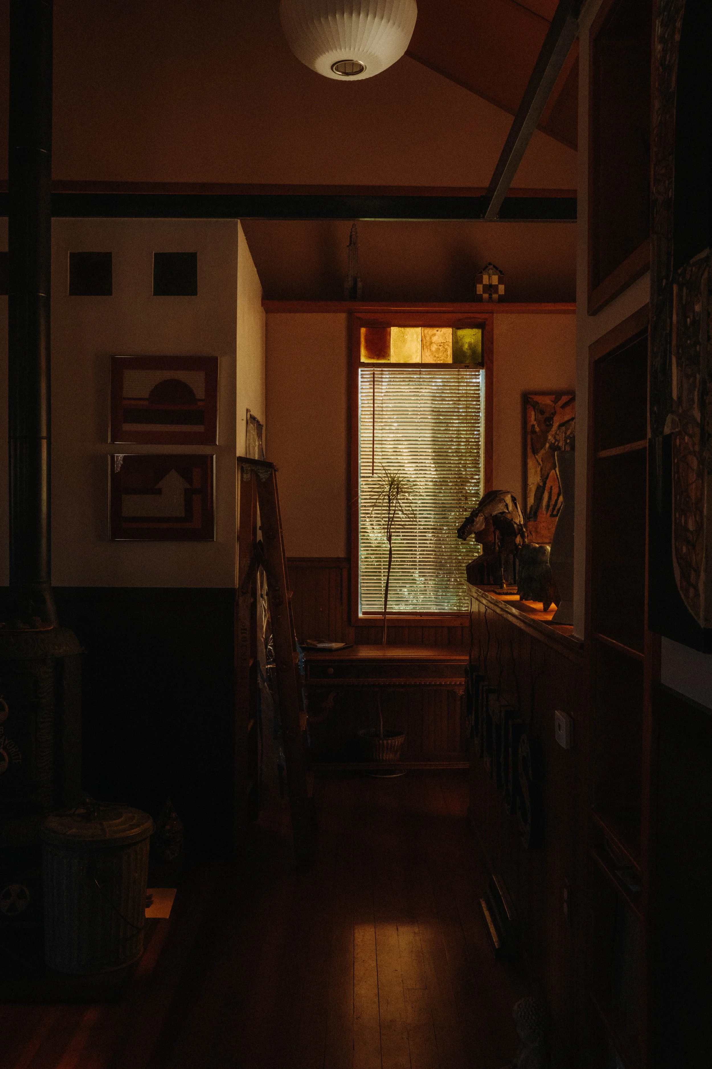 A dimly lit room with a window letting in sunlight, showing blinds and a small tree outside. The room has wooden walls and furniture, with artwork on the walls and various objects, including a ladder and bust sculpture, inside.