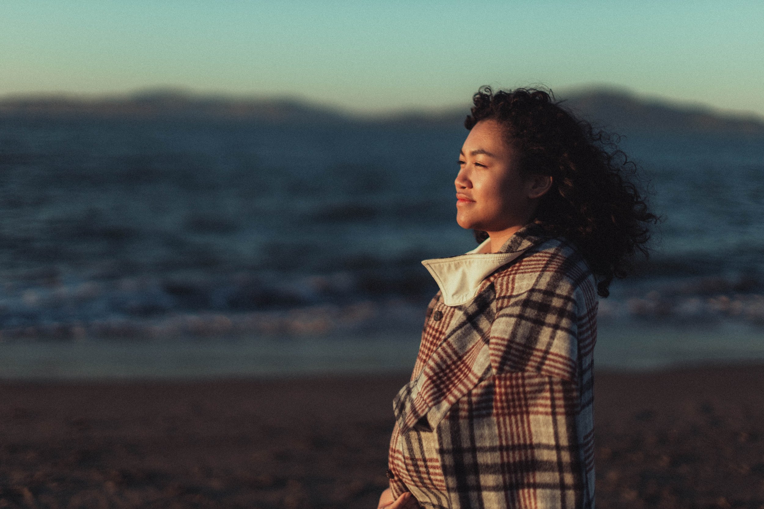 A woman standing on the beach with her eyes closed, facing the ocean, during sunset, wearing a plaid jacket.