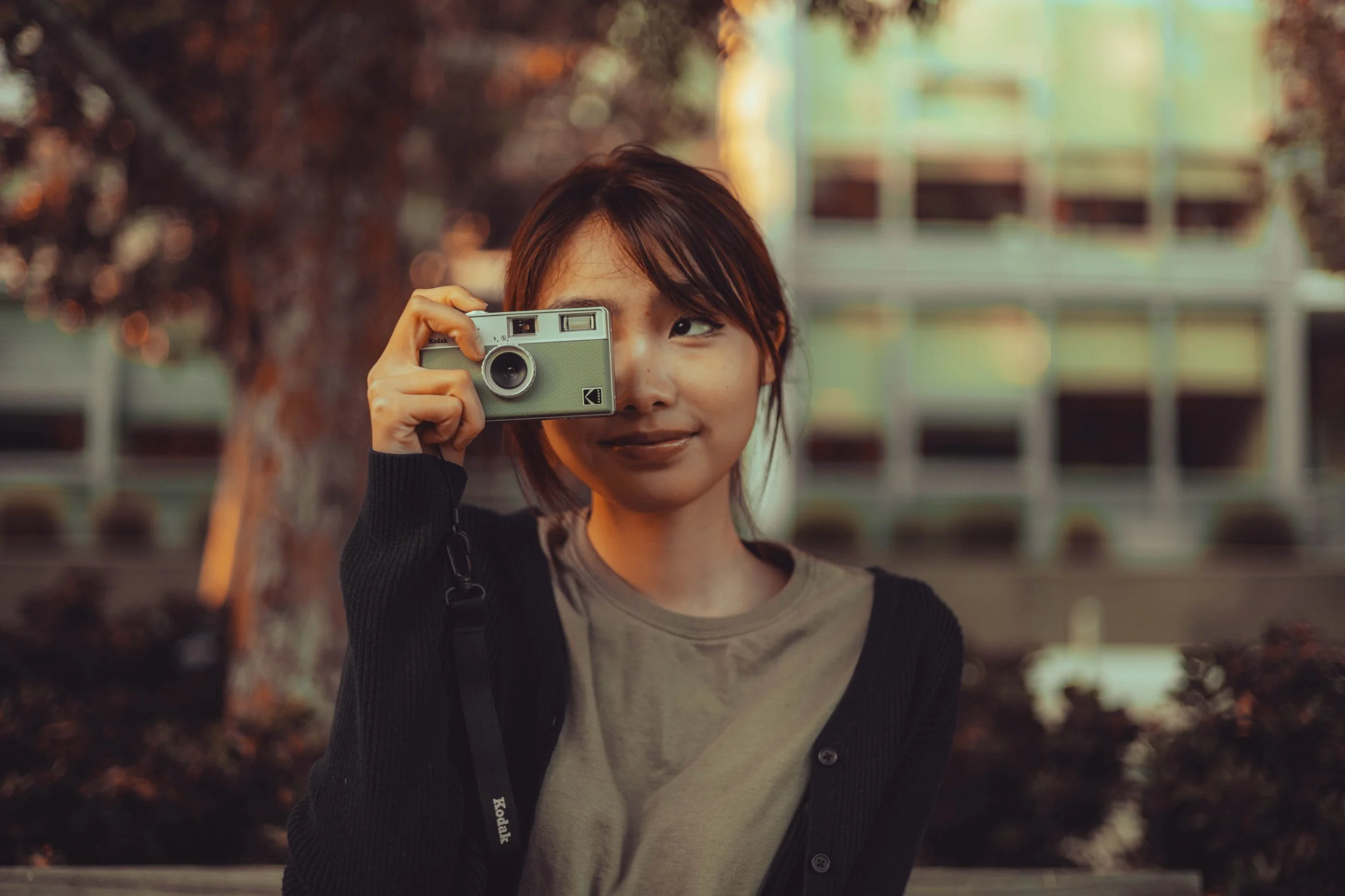 Young woman with short hair takes a photo with a vintage camera outdoors in a park during late afternoon.