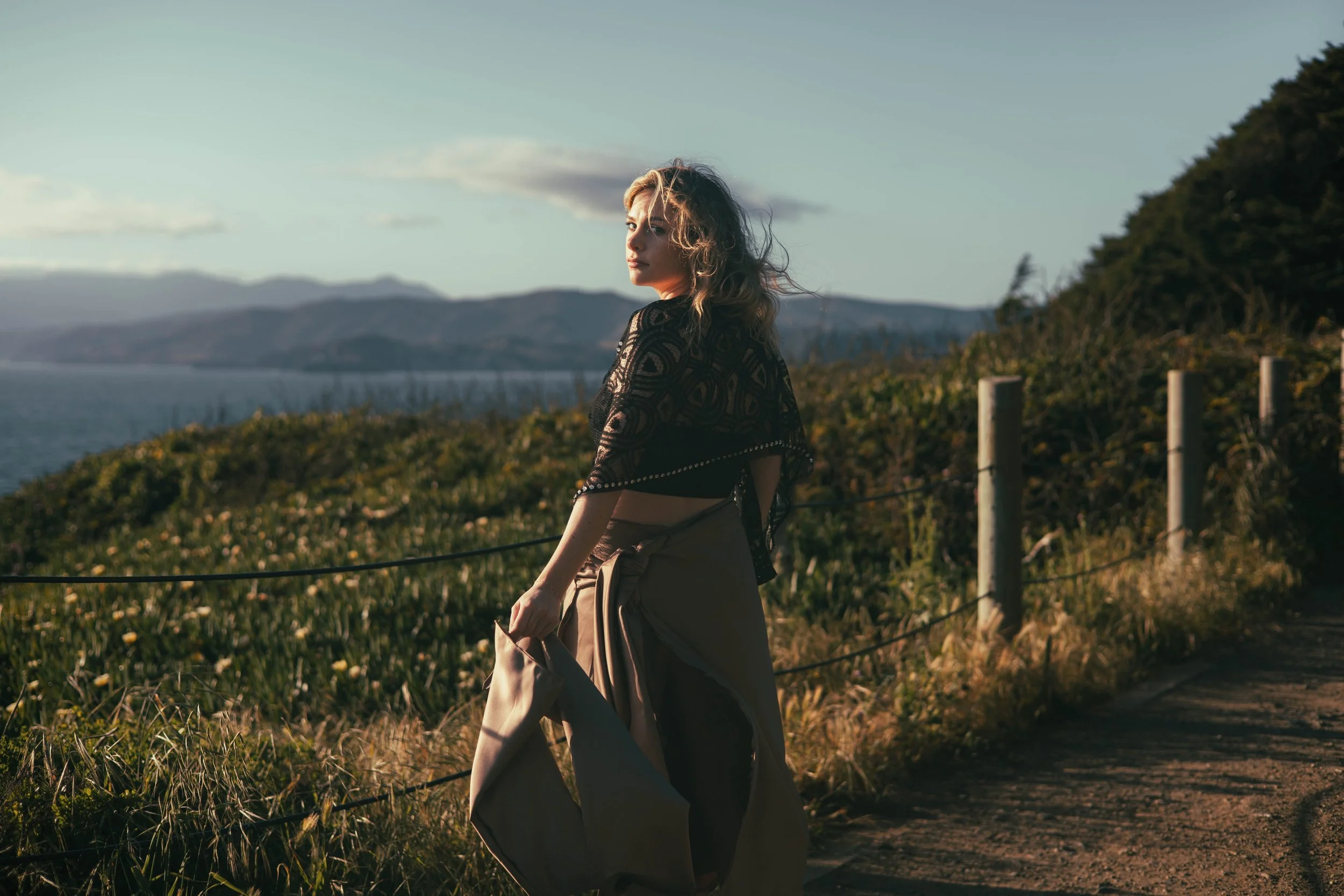 Woman standing on a dirt path near greenery, ocean, and mountains in the distance during sunset.