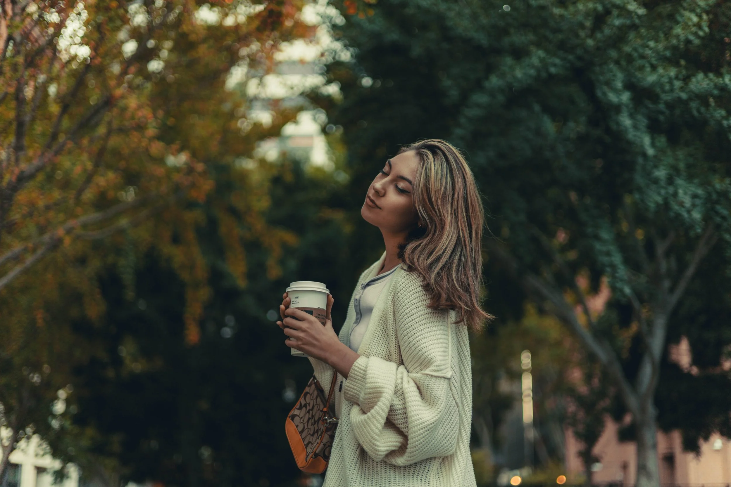 A young woman with wavy light brown hair and fair skin, wearing a cream colored cardigan and white shirt, stands outdoors in a park with trees showing fall foliage, holding a take-out coffee cup, with eyes closed and a content expression.