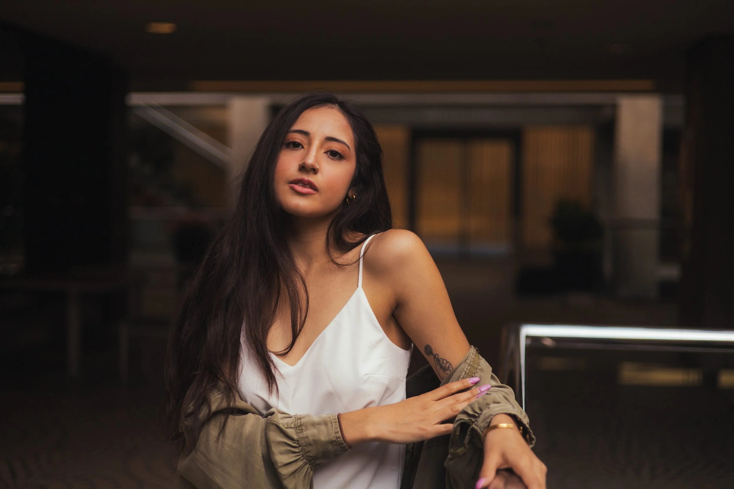 Young woman with long dark hair wearing a white camisole and a beige jacket, posing indoors with a blurred background.