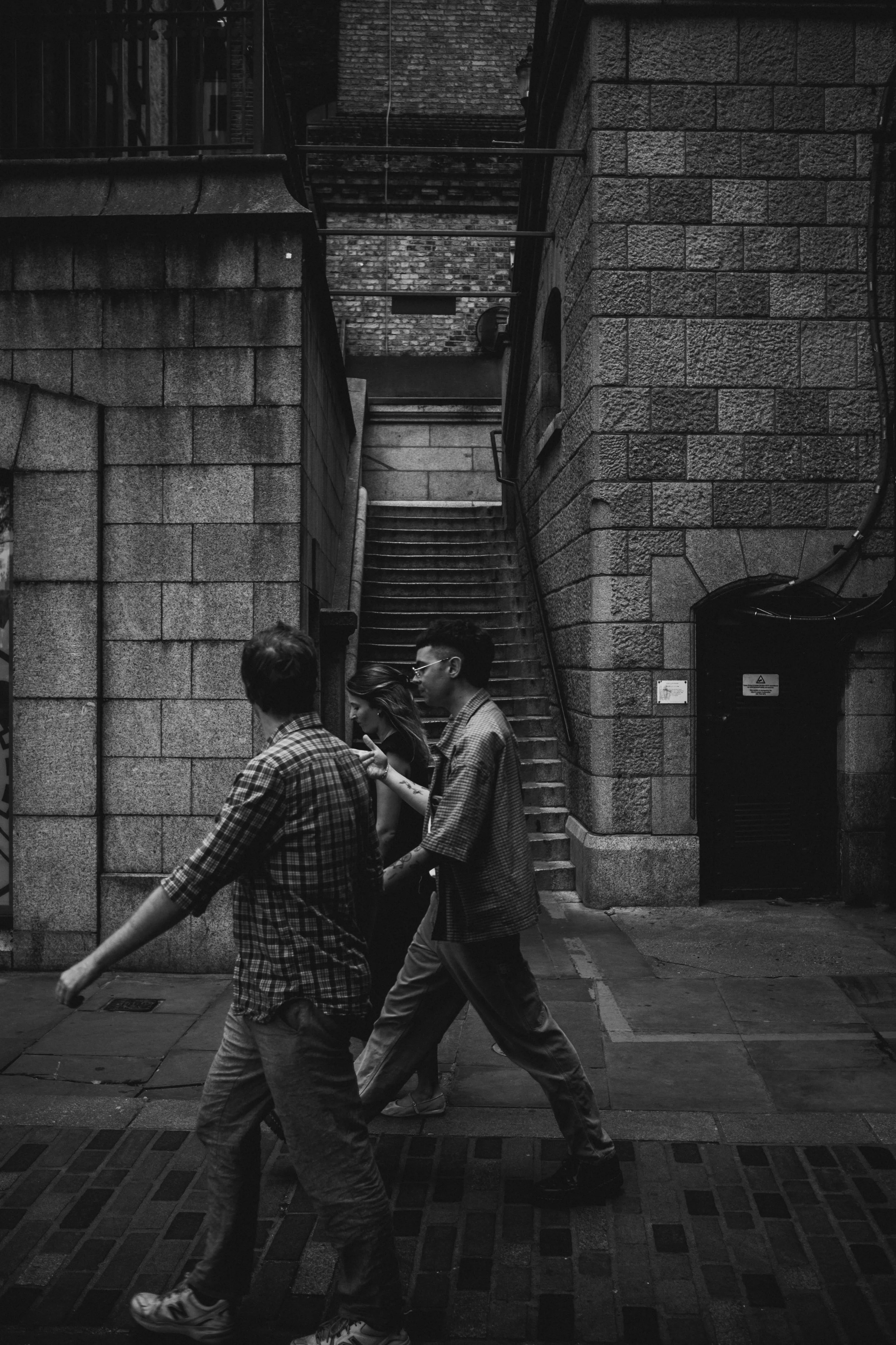 Three people walking across a city street at night, with a staircase and brick building in the background.