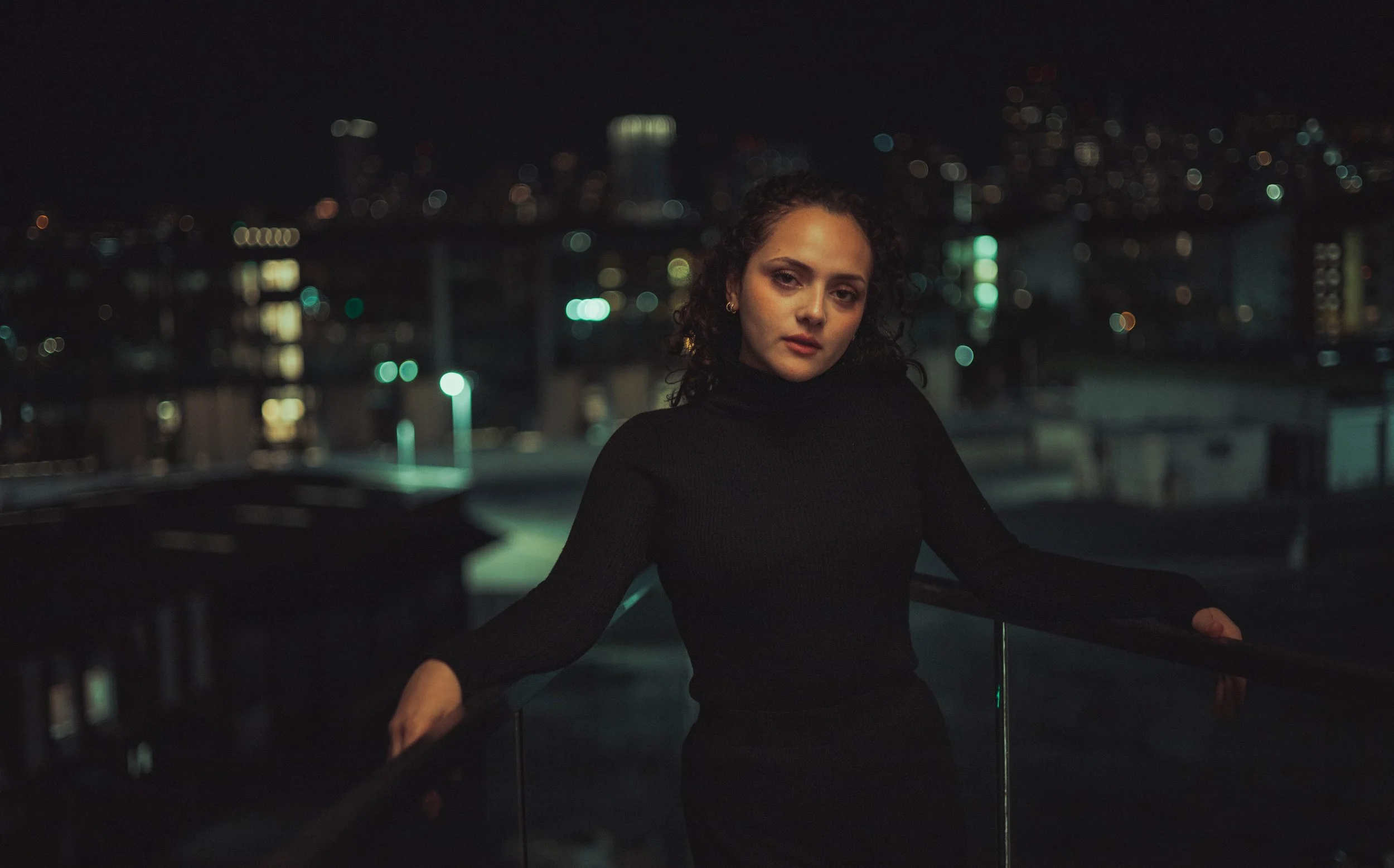 A woman in a black turtleneck sweater standing on a rooftop at night with a city skyline in the background.