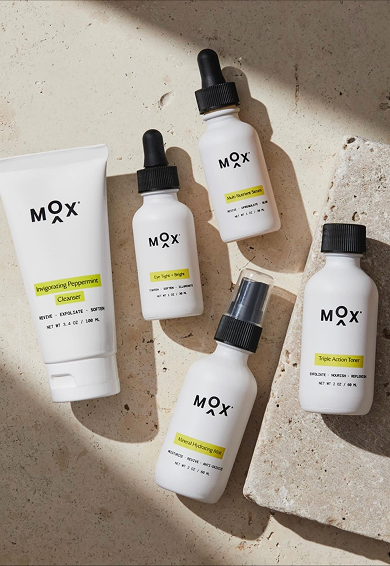 Five skincare products from MOX, including a cleanser, a vitamin serum, a brightening toner, a hydrating mist, and a firming eye gel, arranged on a concrete surface with a rock piece.