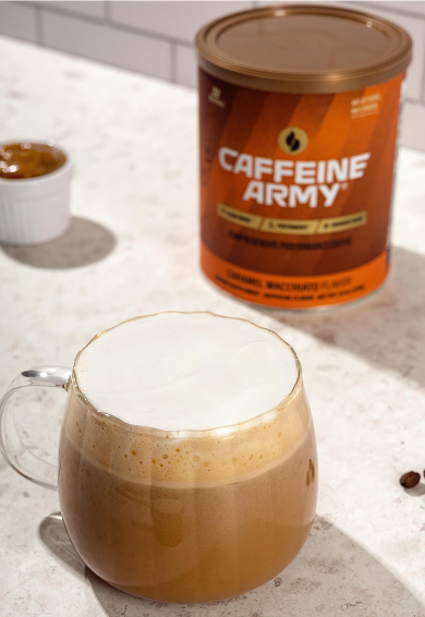 A glass mug filled with a frothy coffee drink topped with whipped cream, with a container of Caffeine Army protein powder and a small dish of caramel in the background.