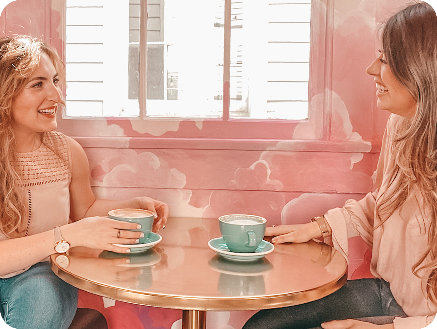Two women sitting at a round table, smiling and chatting, each holding a teal coffee cup on a saucer. They are inside a cafe with pink walls and a large window behind them.