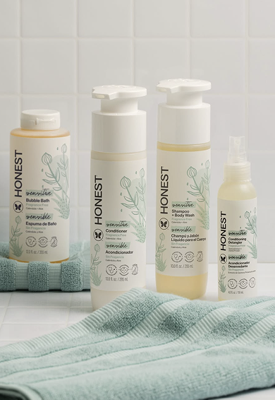Collection of HONEST brand hair and body care products with mint-green towel on white tiled background.