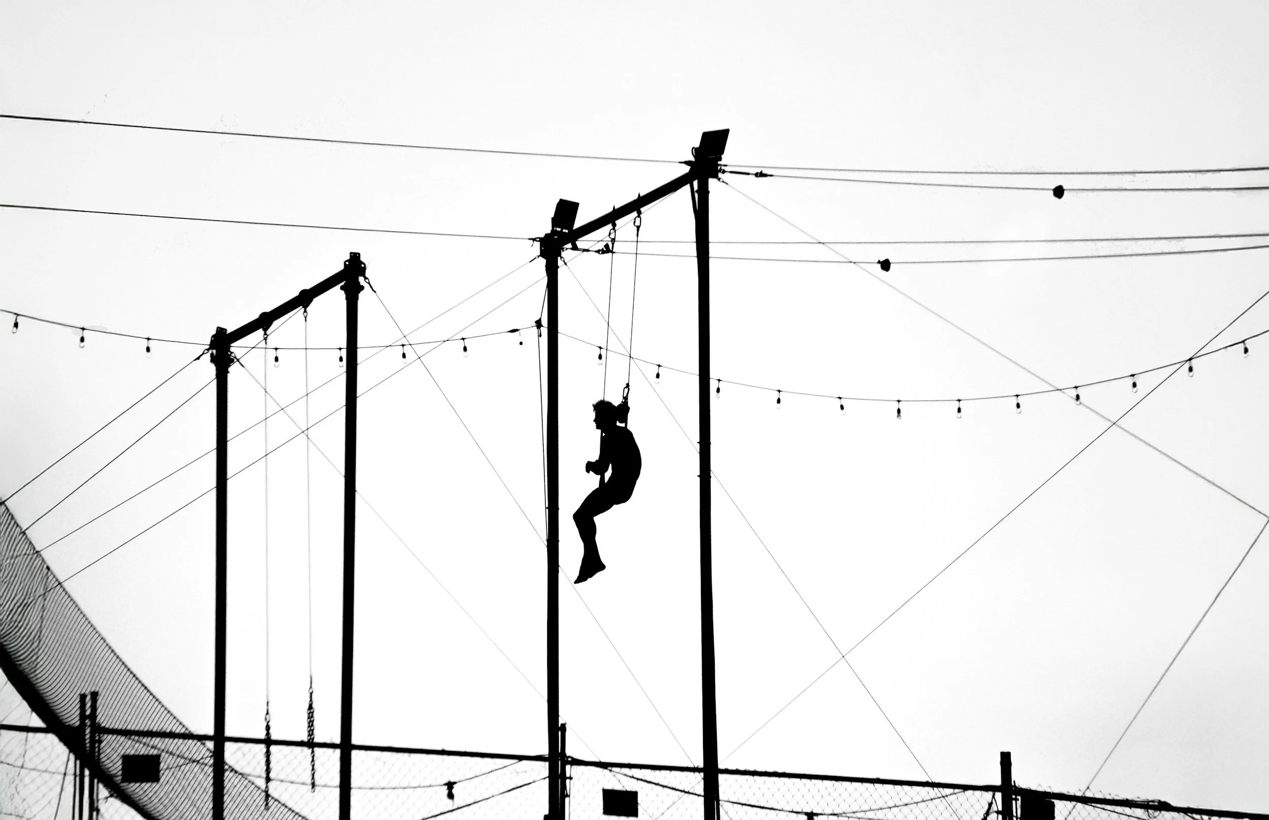 Silhouette of a person sitting on a trapeze bar with multiple wires and string lights in the background.