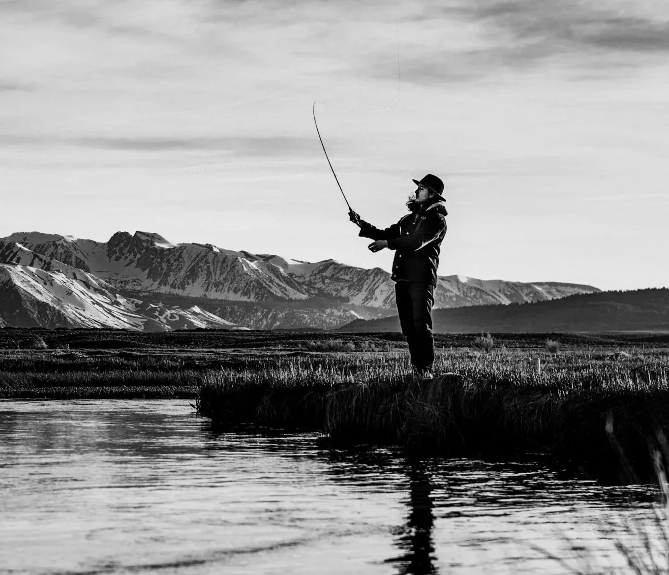 A person standing on a grassy riverbank fishing with a pole, with mountains in the background, in a black and white landscape photograph.