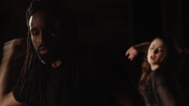 A dark scene featuring two dancers, a man with long dreadlocks in a black shirt in the foreground and a woman with red hair and makeup in the background, both captured in expressive dance poses.