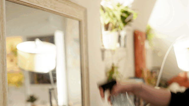 Close-up of a mirror with a person's hand holding a comb or brush, with a blurred background of a room with a lamp, plants, and a window.