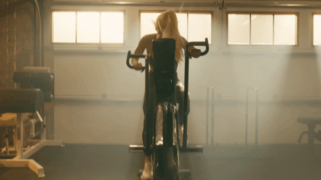A woman working out on an exercise bike in a gym with sunlight coming through the windows.