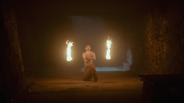 A man performing fire breathing or fire stunts in a dark cave with fire flames in the background.