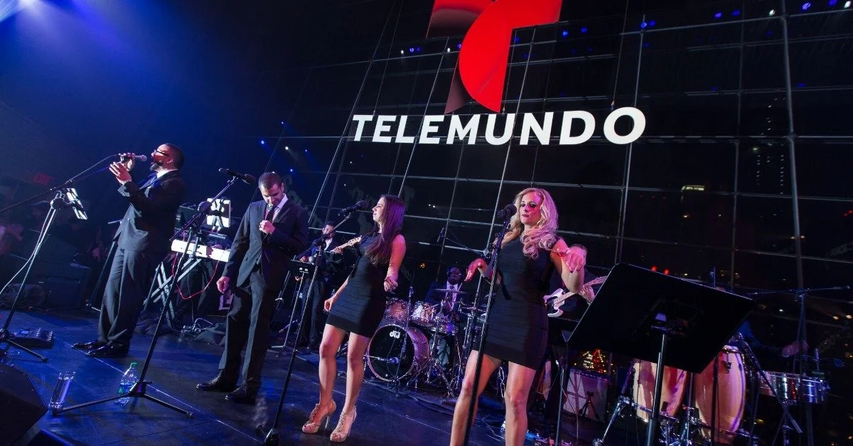A band on a stage with two men and two women at the microphones in front. A sign says "Telemundo" behind them.