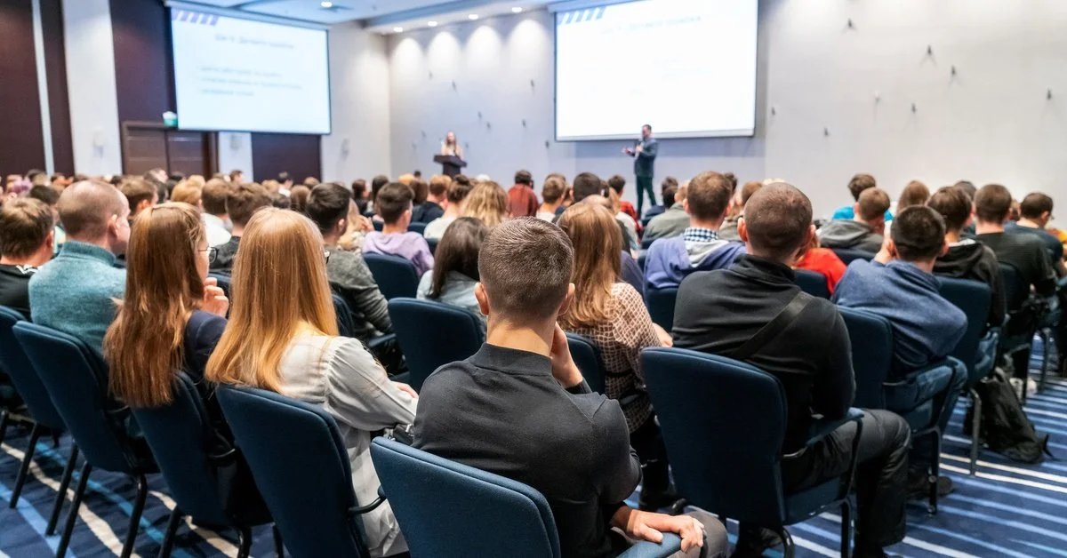 A view from the back of a crowded room with people sitting on chairs watching a business speaker up front.