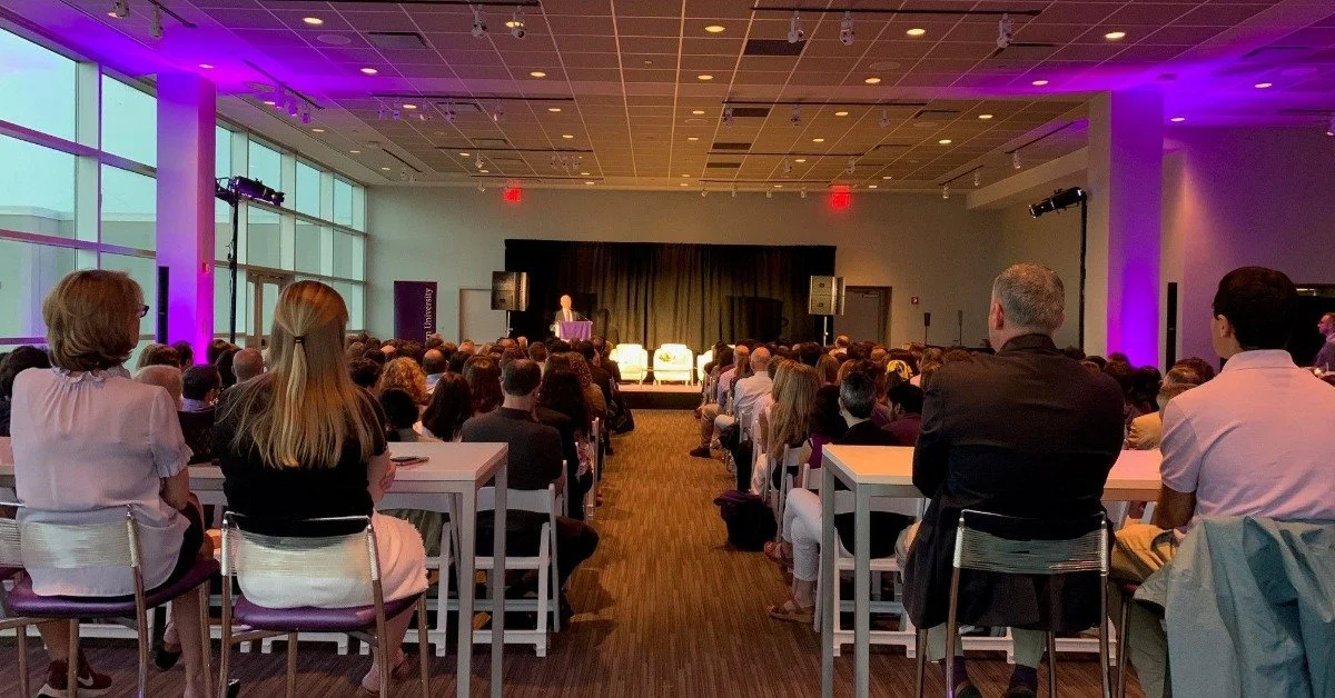 A large conference room full of people all seated and looking at the speaker on the stage in the back.