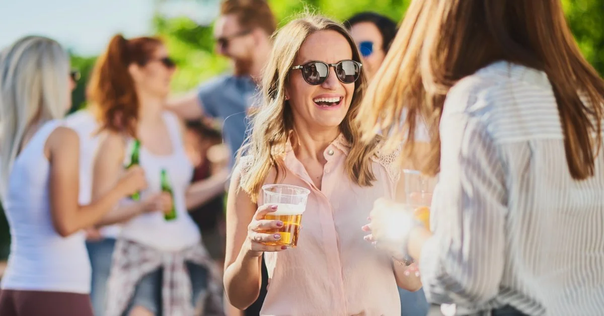 A group of people standing around outside. One woman has a drink in her hand and is laughing with the woman next to her.