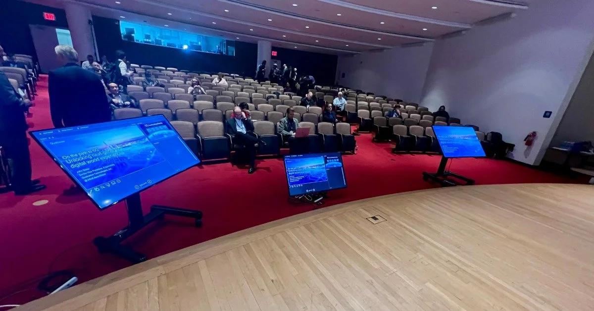 A view from a stage with three computer screens facing the stage and a small crowd in the seats in front of the stage.