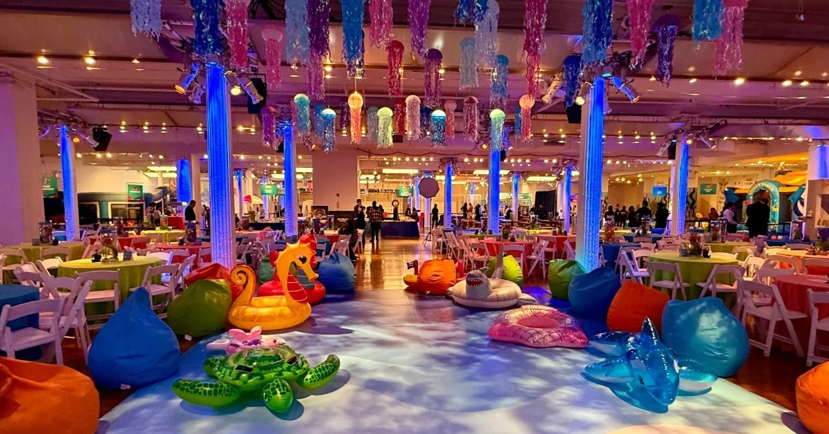 An indoor event venue decorated with an underwater theme, featuring hanging jellyfish decorations, inflatable sea creatures, and banquet tables under vibrant blue and purple lighting.