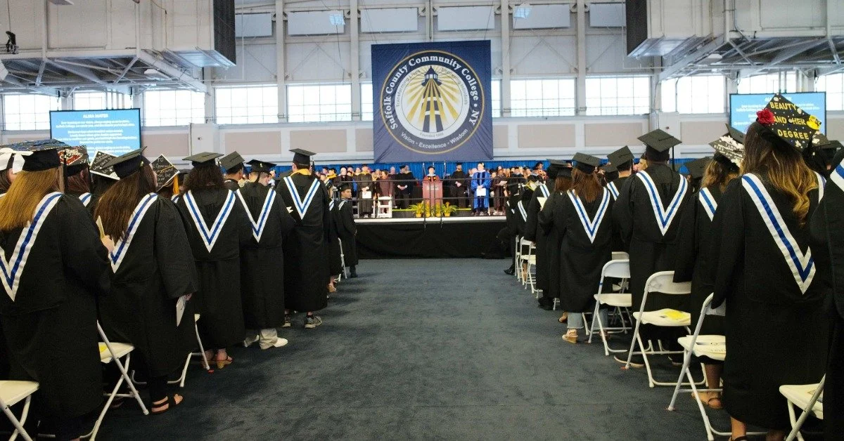 A graduation ceremony taking place indoors. There are many graduates in caps and gowns standing up from their chairs.