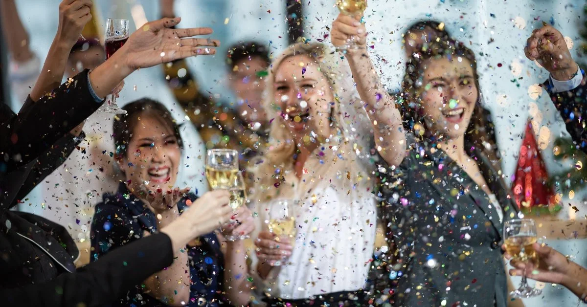 A group of people in nice clothes celebrating together. They are raising glasses as confetti rains down.
