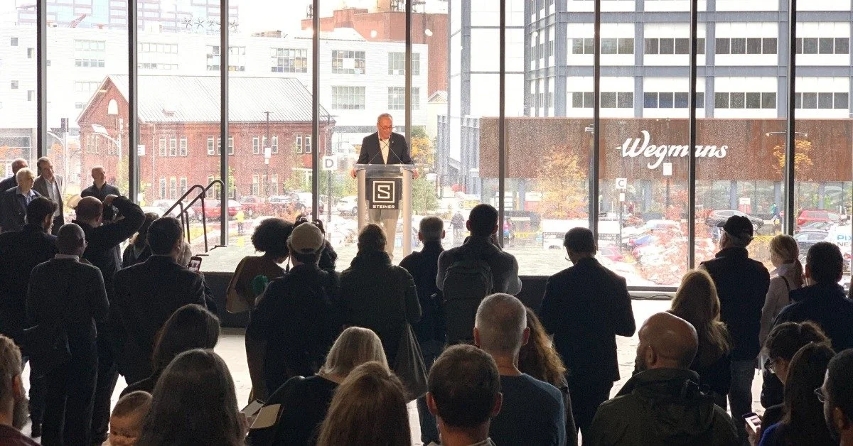 A large group of business people gathered in a crowd standing in front of a man at a podium giving a speech.