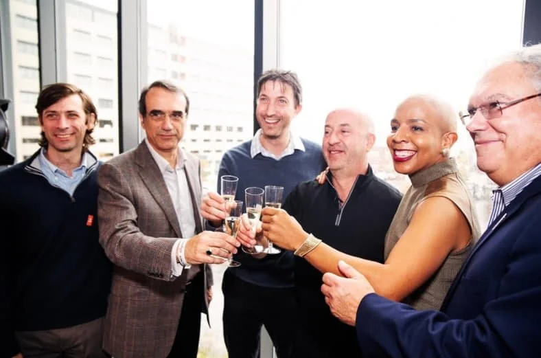 A group of six business professionals in formal attire holding up champagne glasses in a cheers motion.