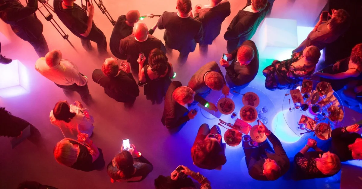 A top-down view of a large corporate event. There are multiple people in business suits gathering around glass tables.