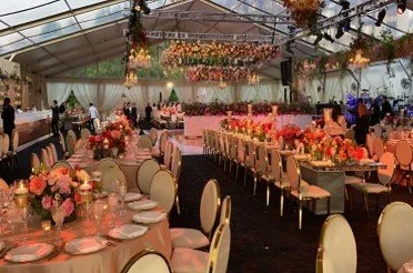 A fancy wedding reception set up inside a glass-roofed building. White chairs surround several large tables.