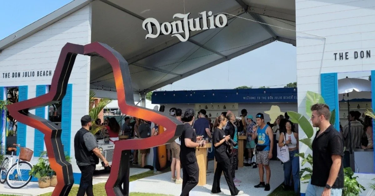 A white event structure with blue shutters, a bar area, people, plants, and a large freeform sculpture.
