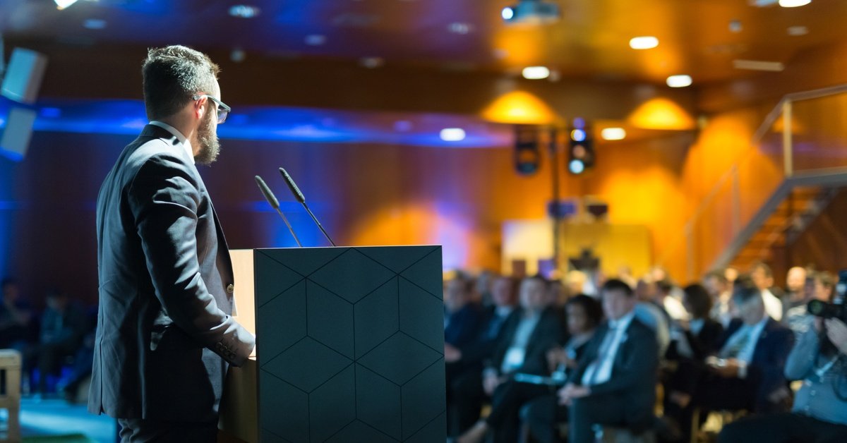 A corporate speaker in professional dress addresses a crowd of attendees in a speech from behind a lectern.