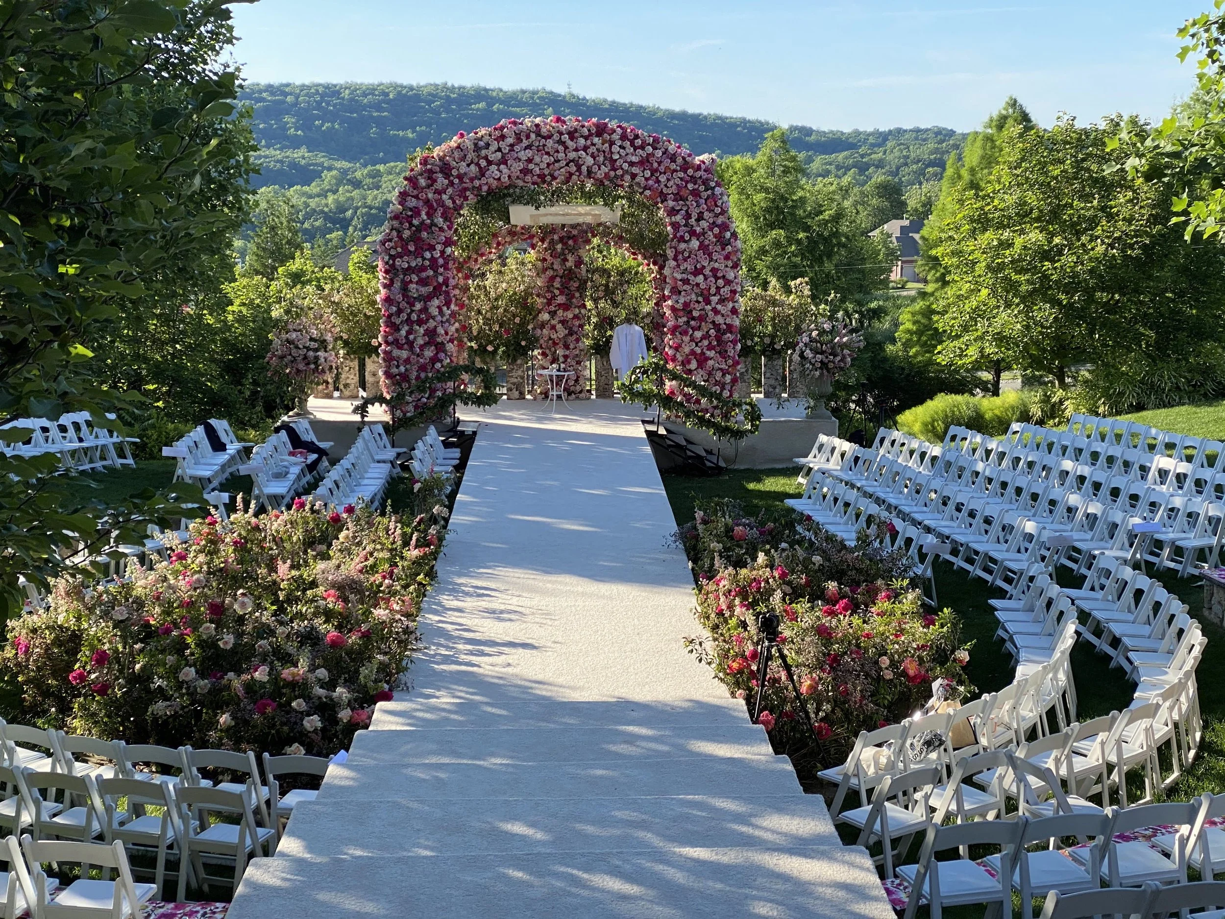 A large outdoor wedding venue with a lot of white chairs all set up in a semi-circle around the wedding archway.