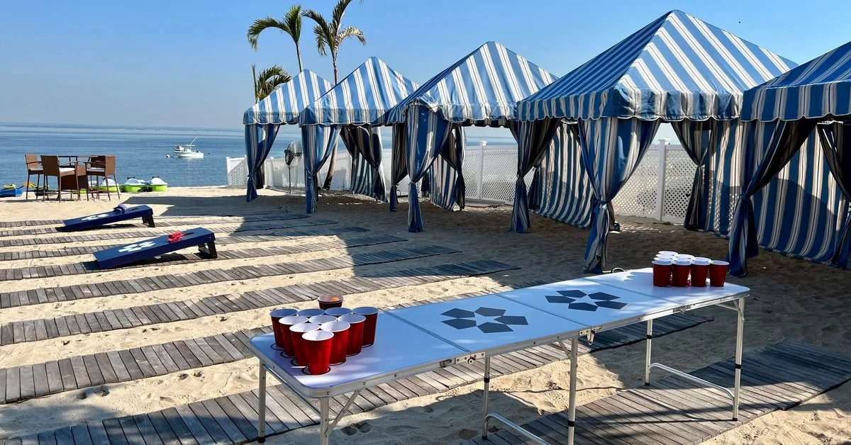 A line of blue and white cabanas on a beach. There are cornhole boards and a beer pong table in front of them.