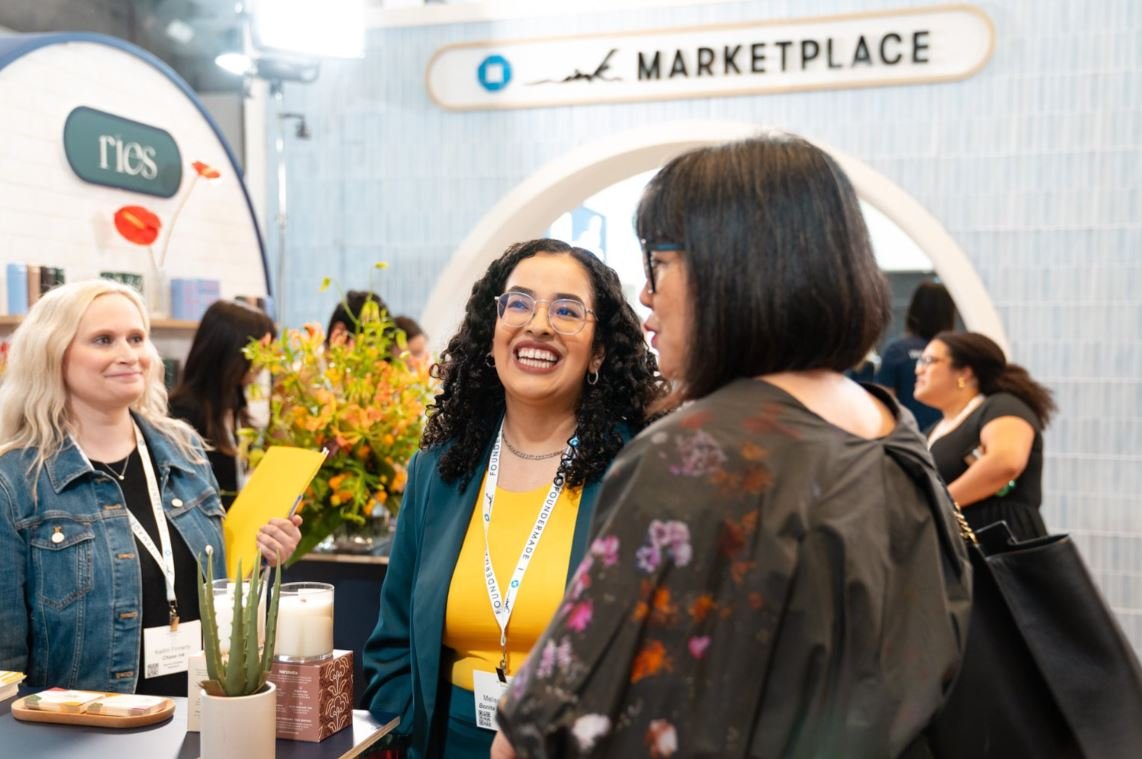 A group of three women at a corporate conference talking to one another over a booth setup under an archway.