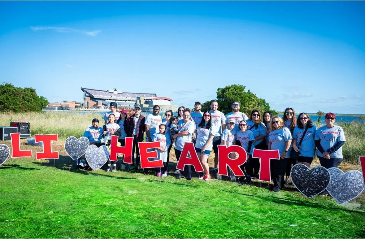 A group of people standing together outside, all wearing the same shirts. There are large, cutout letters in front of them.